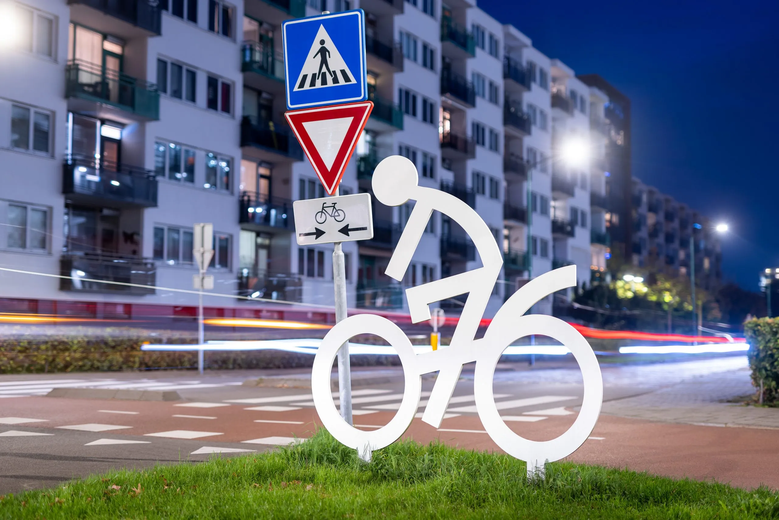 Wit stalen attentiefiguur van een fietser langs een Bredase weg bij avond, met lichtsporen van passerend verkeer