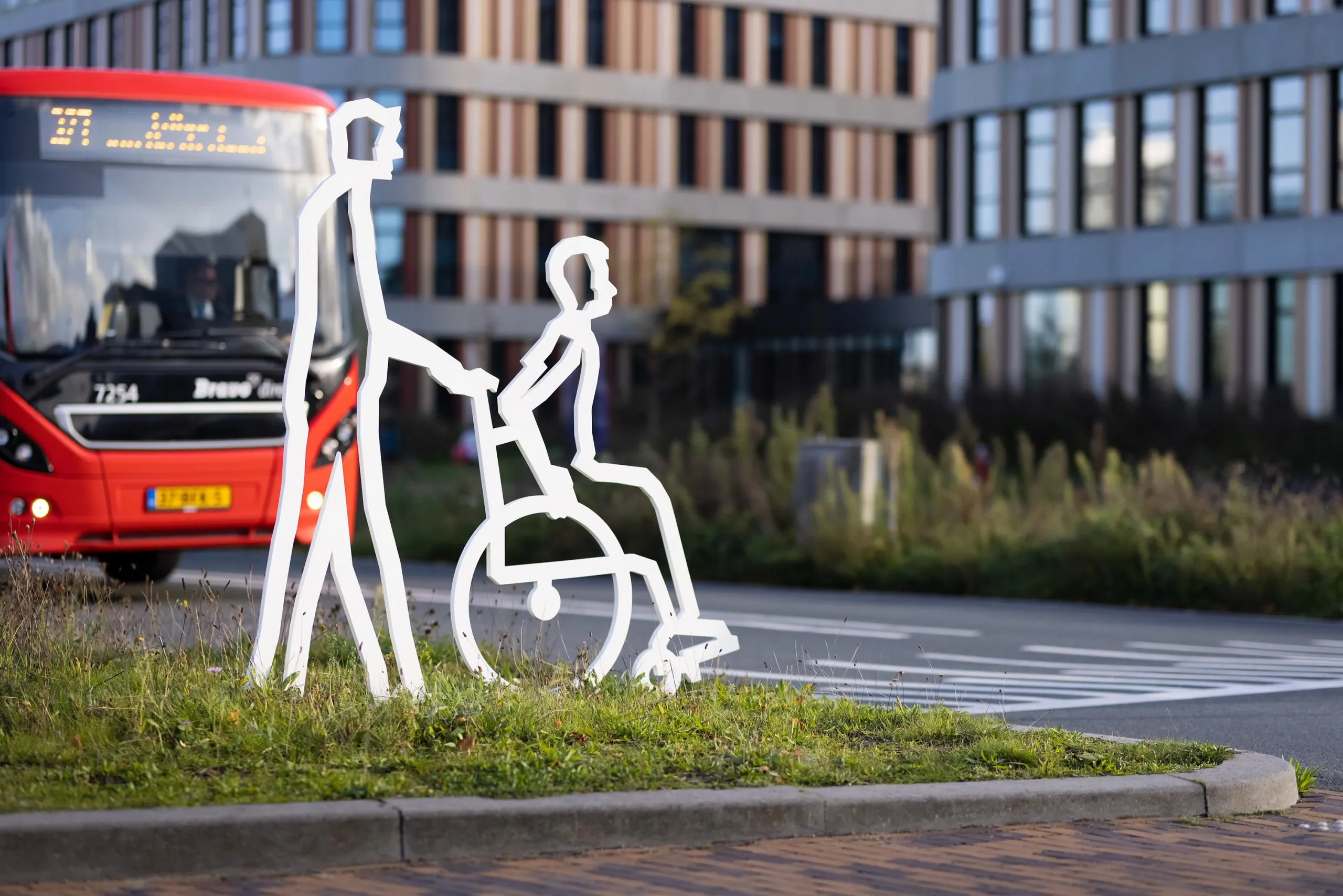 Wit stalen figuur in een rolstoel, met een stadsbus op de achtergrond