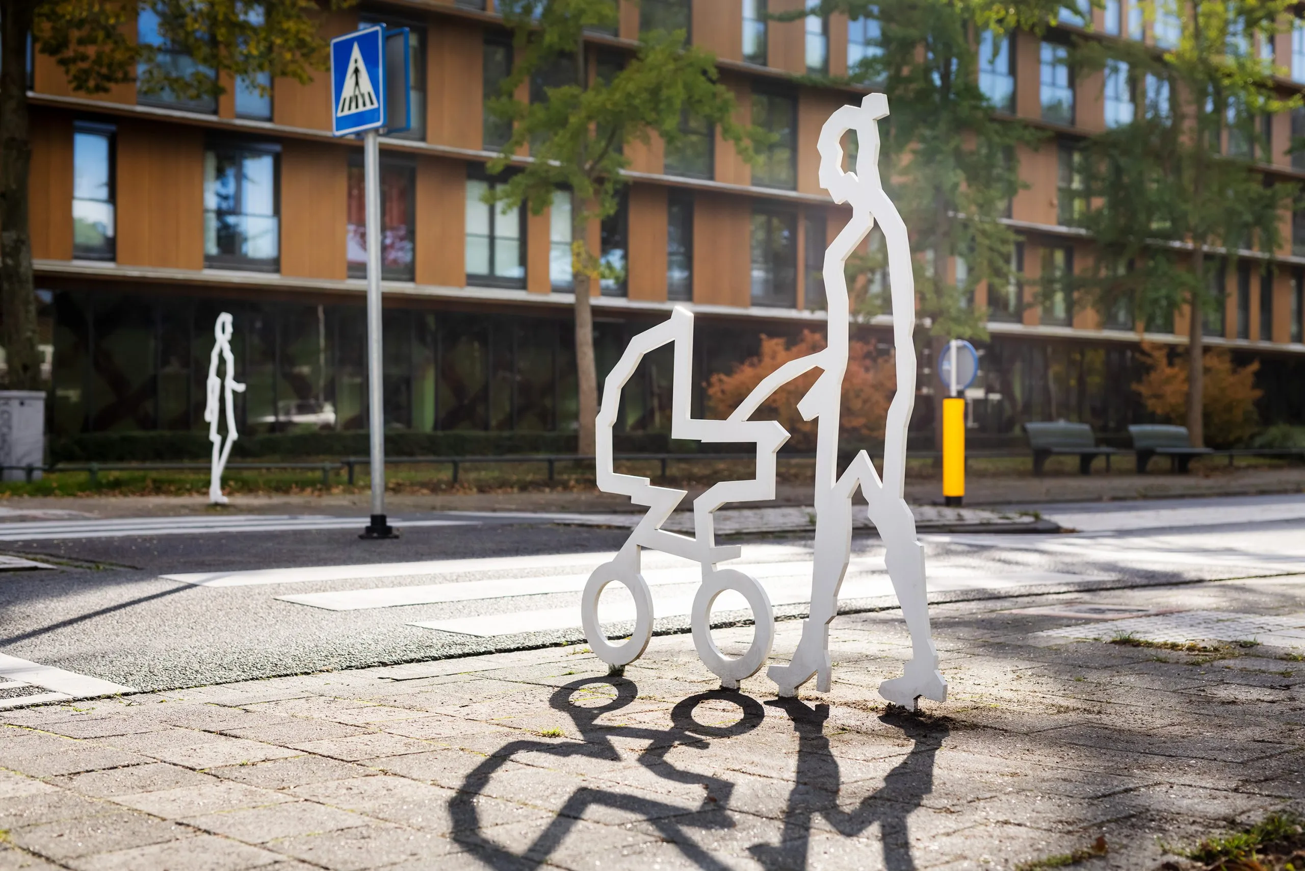 Wit stalen figuur met kinderwagen bij een zebrapad in de ochtendzon, met een tweede figuur op de achtergrond