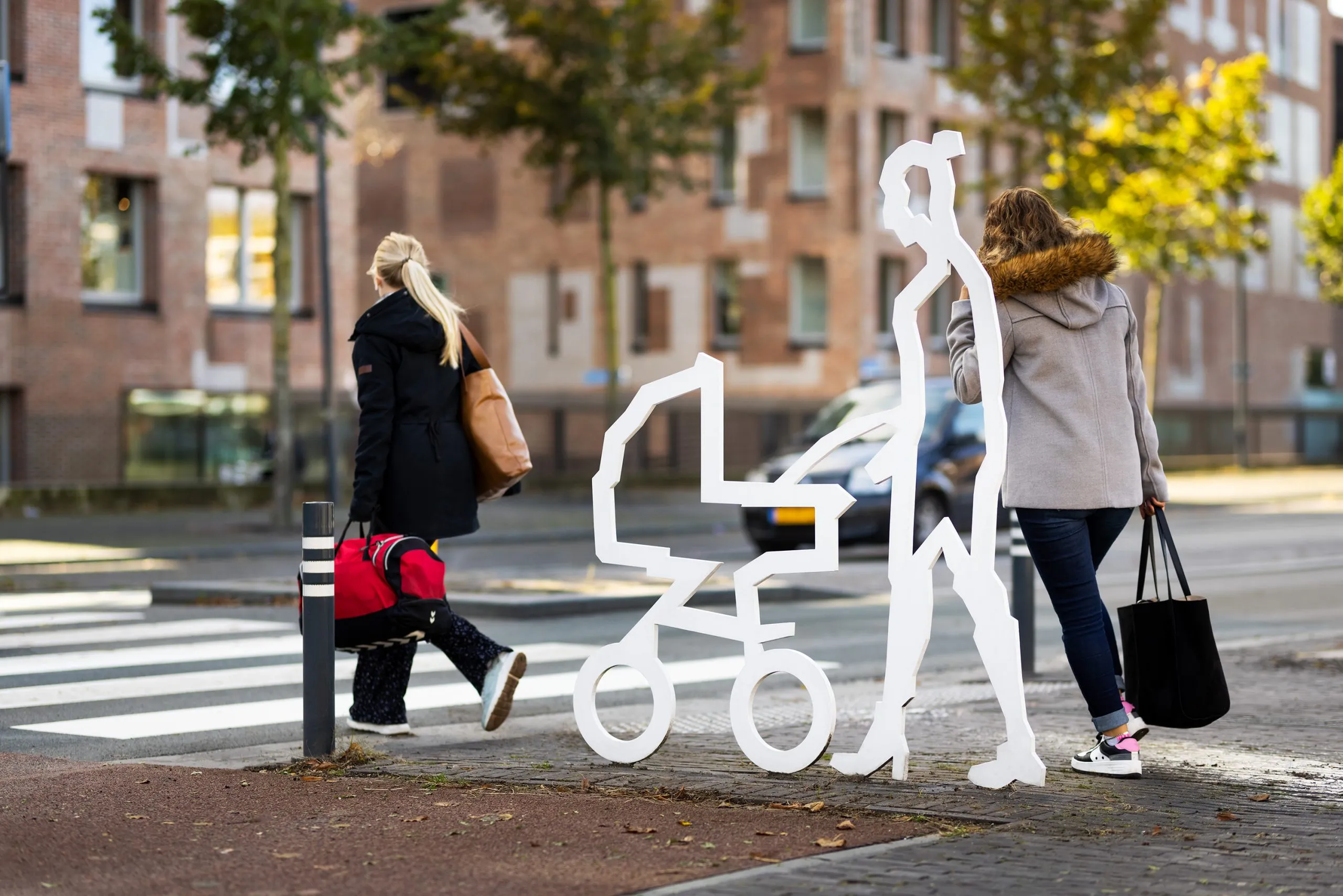 Wit stalen figuur van iemand met een kinderwagen bij een zebrapad in Breda, terwijl echte voetgangers passeren
