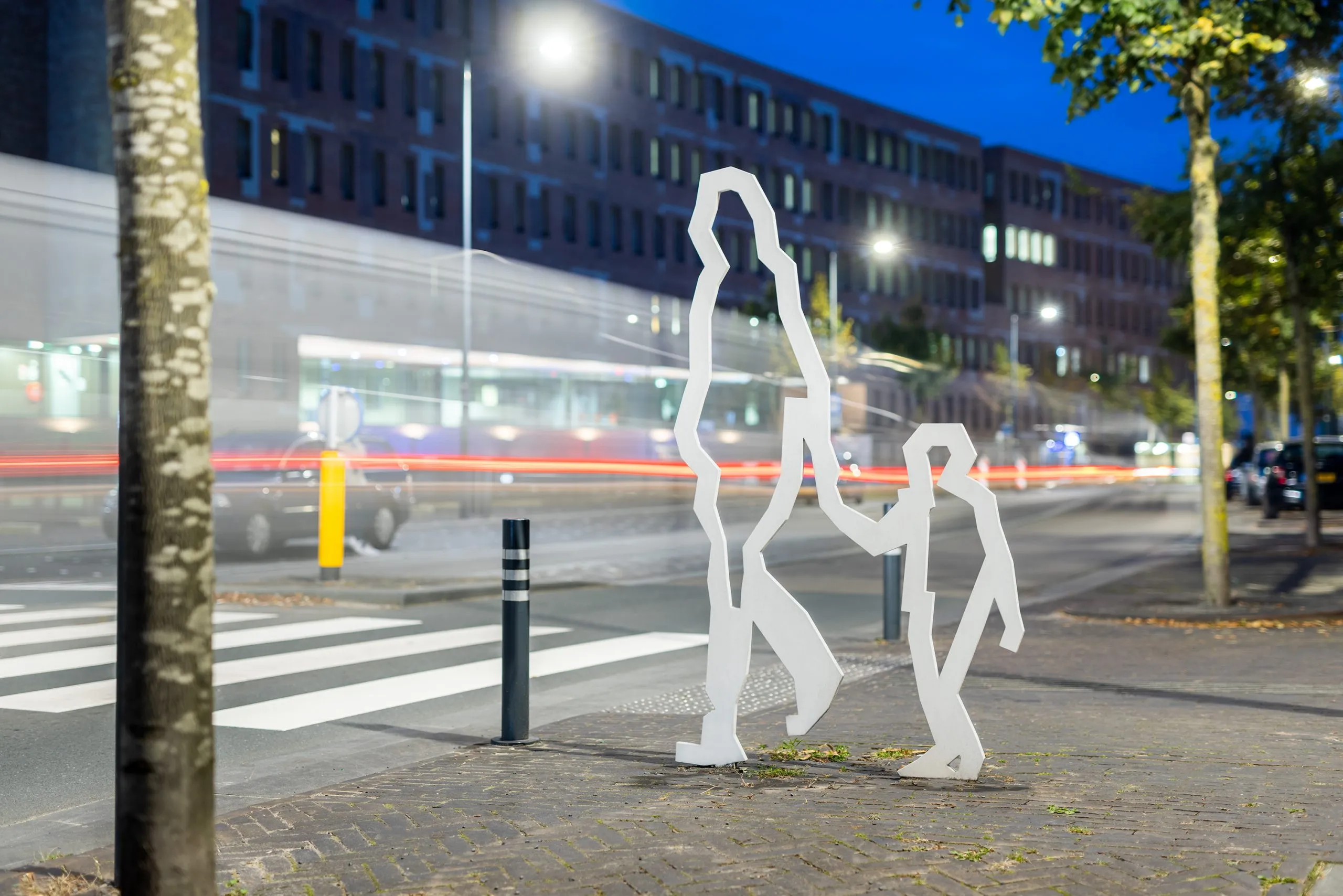Twee witte stalen figuren bij avond, met lichtsporen van passerend verkeer