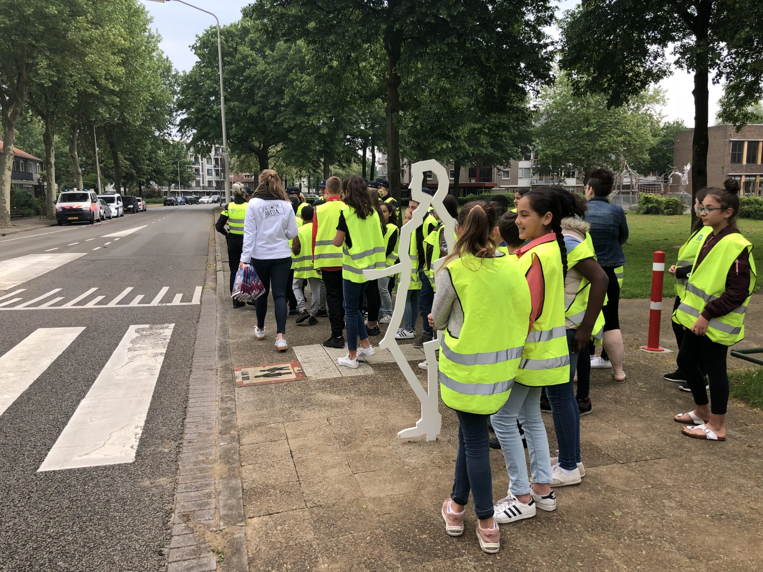 Schoolklas in hesjes bij een oversteek aan de Baliëndijk in Breda, naast een wit stalen figuur